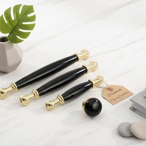 May include: Black and gold cabinet hardware set, including three pull handles and a knob. The handles have a black center and gold accents. A small vase with a green leaf is also present.