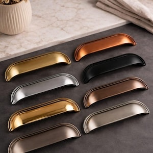 May include: A collection of eight cabinet pulls in various metallic finishes, including gold, silver, black, and bronze. Each pull has a curved, half-moon shape with a detailed edge, arranged on a dark gray surface.