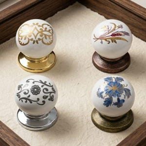 Puede incluir: Cuatro pomos decorativos para armarios con esferas de cerámica blanca y bases metálicas. Cada pomo presenta un diseño diferente: filigrana dorada, ramitas florales, floral gris y floral azul con detalles dorados. Las bases son doradas, bronce, plateadas y latón antiguo.