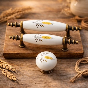 Op de afbeelding: Twee witte keramische kastgrepen en een bijpassende knop. Elke greep en knop heeft een tarwe-ontwerp in geel en bruin, met antieke messing hardware. Op een houten oppervlak.