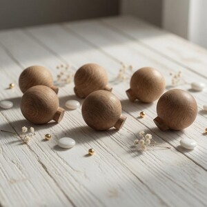 Puede incluir: Seis pomos de madera para armarios con forma redonda y una pequeña base rectangular. Los pomos son de madera de color marrón claro y están dispuestos sobre una superficie de madera blanca, junto con pequeños detalles decorativos blancos y dorados.