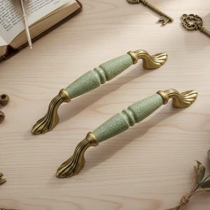 Puede incluir: Dos tiradores decorativos para armarios con un centro verde craquelado y extremos de latón antiguo. Se encuentran sobre una superficie de madera clara, junto con llaves antiguas y un libro, lo que sugiere un tema vintage o antiguo. Miden unos 15 cm.
