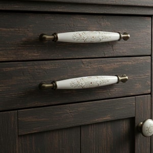 Puede incluir: Primer plano de un mueble de madera marrón oscuro con dos cajones y una puerta. Los cajones tienen tiradores de porcelana blanca con diseño floral y herrajes de latón. La puerta tiene un pomo a juego. El mueble tiene un acabado envejecido.