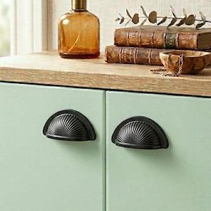 Może przedstawiać: Jasnozielona szafka z dwoma czarnymi uchwytami w kształcie wachlarza. Na górze brązowa szklana butelka ze złotą nakrętką stoi obok stosu książek i małej drewnianej miski. Szafka ma jasny drewniany blat.