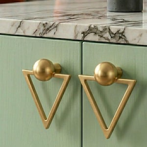May include: Close-up of two cabinet doors with mint green paint and gold hardware. Each door has a gold triangle-shaped handle with a spherical knob. The handles are mounted on the cabinet doors. The cabinet is under a marble countertop.