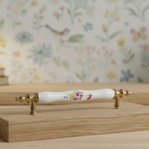May include: A cabinet pull with a white ceramic handle decorated with flowers. The pull has gold-coloured metal ends and is mounted on a wooden block. The design is reminiscent of vintage styles.