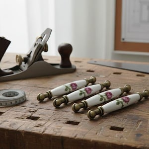 May include: A collection of woodworking tools and hardware on a wooden surface. Includes a hand plane, measuring tape, a metal ruler, and four white and gold cabinet pulls with a floral design.