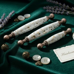 May include: White and gold speckled cabinet hardware with bronze-colored accents. The set includes three pulls and one knob, along with decorative buttons and metal filigree balls. The text on the card reads "Heirloom Collection."
