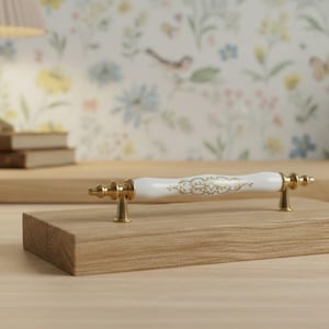 May include: A white and gold cabinet pull with a floral design is mounted on a small wooden block. The pull has a white ceramic center with gold accents. The wooden block is light brown. The background includes a lamp, books, and a vase.