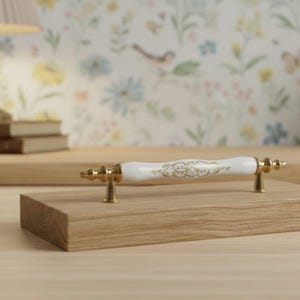 May include: A drawer pull featuring a white ceramic handle with a gold filigree pattern, accented by polished gold metal end caps and mounting posts, mounted horizontally on a rectangular piece of light-toned wood with visible grain.