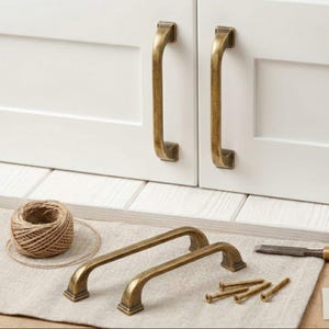 May include: Antique brass cabinet pulls and screws are displayed with a roll of twine. Two pulls are attached to a white cabinet door. The pulls have a curved design and square base. The hardware is for home improvement projects.