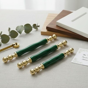 Op de afbeelding: Drie groene en gouden kastknoppen met een bijpassend meetinstrument en een takje groen. De knoppen hebben een donkergroen midden en gouden accenten. Een witte kastdeur en houtmonsters staan op de achtergrond.