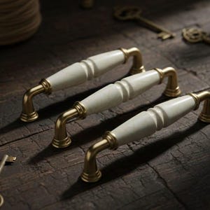 May include: Three cabinet pulls with white ceramic centers and gold-colored metal hardware. The pulls are arranged on a dark wooden surface, with antique keys and decorative items in the background. The hardware is a classic design.