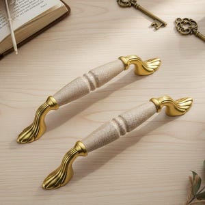 Könnte beinhalten: Zwei Schrankgriffe mit cremefarbenem Mittelteil und goldfarbenen, verzierten Enden. Die Griffe liegen auf einer hellen Holzoberfläche, zusammen mit antiken Schlüsseln, einem offenen Buch und dekorativen Elementen.