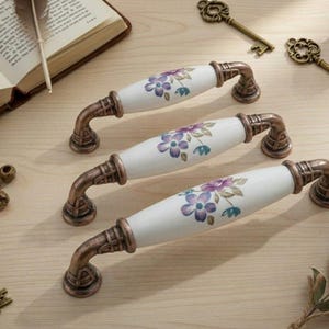 May include: Three decorative cabinet pulls with white ceramic handles adorned with purple and blue floral patterns. The pulls have bronze-colored metal bases and curved arms. The image also shows antique keys and a book.