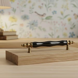 Op de afbeelding: Een zwarte en gouden kastgreep is gemonteerd op een klein, lichtgekleurd houten blok. De greep heeft een glanzend zwart midden en goudkleurige accenten. De greep is een decoratief element voor meubels.