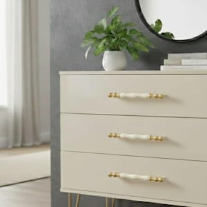 May include: A cream-coloured three-drawer chest of drawers with gold-toned legs and handles. The handles are white with gold accents. A small potted plant sits on top, near a round black mirror.