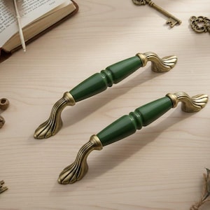 May include: Two dark green and gold cabinet handles with a vintage aesthetic. The handles are placed on a light wood surface, along with antique keys, beads, and an open book. The handles have a classic, ornate design.