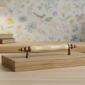 Puede incluir: Un tirador de mueble con una barra central de cerámica color crema lisa y forma ondulada. El tirador está sujeto por dos soportes metálicos de tono bronce con un diseño decorativo escalonado a cada lado de la empuñadura central.