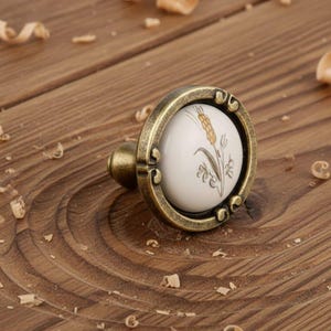 May include: A decorative cabinet knob with a white ceramic centre featuring a wheat stalk design. The knob has a bronze-coloured metal frame and a small, rounded handle. The background is a wooden surface.