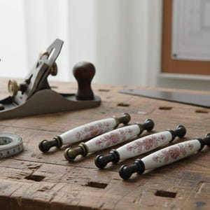May include: A woodworking scene with a hand plane, measuring tape, and four decorative cabinet pulls. The pulls are white with a floral design and black and brass accents. The items are on a wooden workbench.