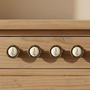 Op de afbeelding: Vier antieke messing kastknoppen met witte keramische centra. Elke knop heeft een tarwe-aar ontwerp in geel en groen. De knoppen zijn gemonteerd op een lichtbruin houten oppervlak.