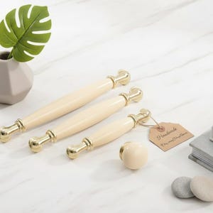 Peut inclure: Tiroirs de meubles crème et bouton avec des accents dorés. Le matériel est disposé sur une surface en marbre blanc, à côté d'un petit vase avec une feuille verte, une étiquette "Handmade" et une pile de livres.