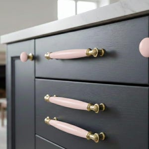 Può includere: Primo piano di un mobile da cucina grigio scuro con ferramenta rosa e oro. L'armadio ha tre cassetti con maniglie rosa e oro e un pomello rosa. Il piano di lavoro è bianco.