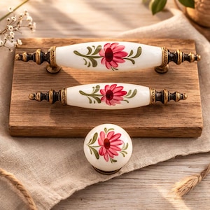 May include: A set of three decorative cabinet hardware pieces: two white ceramic handles and a matching knob. Each piece features a hand-painted pink flower design with green leaves and gold-toned metal accents. The hardware is displayed on a wooden board.