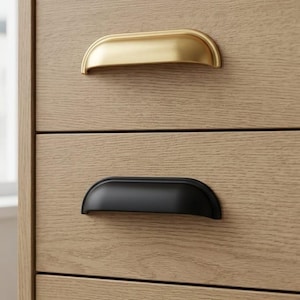 Może przedstawiać: Zbliżenie drewnianej komody z trzema różnymi uchwytami szuflad. Górny uchwyt jest złoty, środkowy czarny, a dolny srebrny. Uchwyty mają kształt półksiężyca.