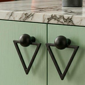 May include: Close-up of a mint green cabinet with two black triangular drawer pulls. Each pull has a spherical knob above a triangular frame. The cabinet has a marble countertop and a small gray vase with a plant.