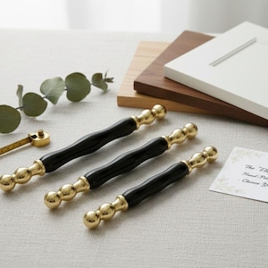 Op de afbeelding: Drie zwarte en gouden kastgrepen met een gebogen ontwerp worden gepresenteerd op een neutrale ondergrond. Een kleine gouden meetlint en een takje groen zijn ook aanwezig. Een witte kastdeur en houten stalen staan op de achtergrond. Een kaart leest "The Elegance Collection".