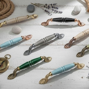 Op de afbeelding: Een verzameling decoratieve kastgrepen in verschillende kleuren, waaronder beige, zwart, blauw, groen en grijs. De handgrepen hebben sierlijke, gebogen ontwerpen met goudkleurige accenten. Een kompas en decoratieve elementen zijn ook aanwezig.