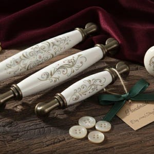 Op de afbeelding: Een verzameling antieke meubelbeslag, waaronder drie witte en gouden ladetrekker, een bijpassende knop en knopen. Het beslag heeft sierlijke bloemmotieven en bronzen metalen accenten. Een sleutel en veer zijn ook aanwezig.