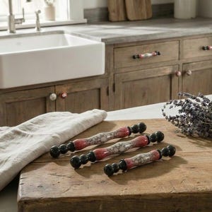 Pode incluir: Uma cena de cozinha rústica com uma tábua de cortar de madeira e três puxadores decorativos para armários. Os puxadores têm detalhes vermelhos e brancos e extremidades pretas. Um ramo de lavanda e uma pequena planta em vaso também estão presentes.