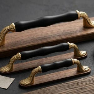 Peut inclure: Trois poignées de meubles avec un centre noir et des extrémités incurvées dorées sont présentées sur des planches de bois. Les poignées sont présentées avec une petite coupelle contenant de la quincaillerie, suggérant un thème de mobilier ou de décoration intérieure.