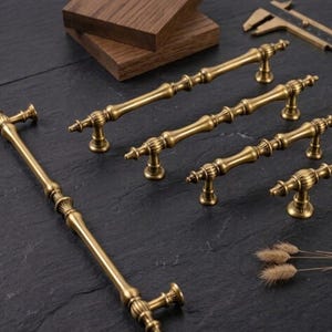 Könnte beinhalten: Eine Sammlung antiker Messing-Schrankgriffe und -Knöpfe. Die Hardware zeichnet sich durch ein detailliertes, verziertes Design mit warmer, goldener Oberfläche aus. Verschiedene Größen werden gezeigt, darunter ein langer Griff und mehrere kleinere Knöpfe.