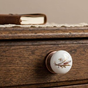 Op de afbeelding: Een houten lade met een witte keramische knop met een goud- en bruin bloemmotief. Bovenop ligt een bruin leren notitieboek. De knop is bevestigd met een koperkleurige metalen plaat.