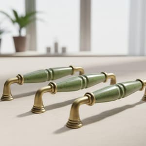 Könnte beinhalten: Drei Schrankgriffe mit grün gesprenkelten Keramikgriffen und Messingfarbenen Metallbeschlägen. Die Griffe sind auf einer hellen Oberfläche angeordnet, passend für Küche oder Möbel.