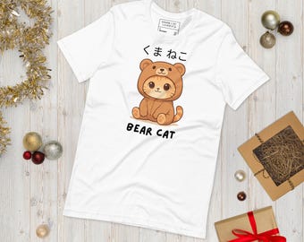Pode incluir: Camiseta branca com um personagem de desenho animado de gato-urso. O gato-urso é marrom e está usando uma fantasia de urso. O texto japonês "くまねこ" e o texto em inglês "BEAR CAT" são impressos acima e abaixo do personagem.