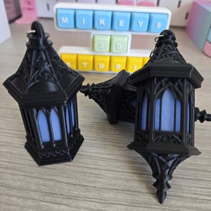 May include: Three black decorative lanterns with a gothic design. The lanterns have a pointed base and a detailed top with a blue light inside. The lanterns are resting on a light-colored wooden surface.
