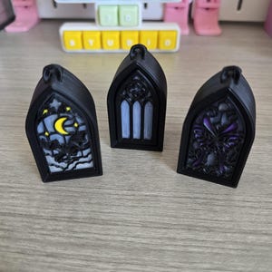 Pode incluir: Três ornamentos pretos em forma de janela gótica. Um apresenta uma lua e morcegos, outro um design de vitral e o terceiro uma borboleta. Cada um tem uma argola para pendurar. O fundo inclui um teclado com o texto "MIKEYS STUDIO".