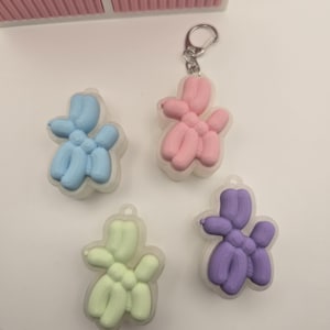 Puede incluir: Cuatro llaveros de perro globo de colores en azul, rosa, verde y morado. Cada llavero está encerrado en un marco de plástico transparente y sujeto a un llavero plateado. Los llaveros están dispuestos sobre una superficie blanca.