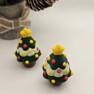 May include: Two miniature Christmas trees with a dark green base, white middle section, and yellow star on top. The trees are decorated with red and yellow dots. A pine cone and a small potted evergreen tree are in the background.