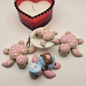 Peut inclure: Une bougie rouge en forme de cœur avec un intérieur en cire blanche est placée au-dessus de plusieurs figurines de tortues fantaisistes. Les tortues sont principalement roses et beiges, avec des vermicelles blancs et divers éléments décoratifs. Une tortue a un porte-clés attaché.