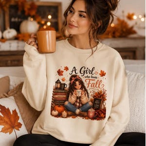 Peut inclure: Un sweat-shirt à col rond couleur crème avec une illustration graphique représentant une personne assise parmi des livres, des citrouilles et des feuilles d'automne. Le texte "A Girl who loves Fall" est imprimé au-dessus de l'illustration.