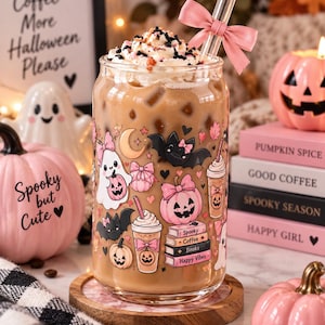 Puede incluir: Un vaso transparente lleno de café helado, cubierto con nata montada y virutas, con un lazo rosa en una pajita de cristal. El vaso está decorado con ilustraciones coloridas de fantasmas, murciélagos, calabazas y libros con texto.