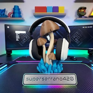 May include: A headset stand featuring a cluster of brown mushrooms with white stems and a blue crystal base. The stand holds a white and black headset. A silver nameplate at the base reads "superserrano420".