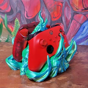May include: A red and maroon Xbox controller set in a teal and blue flame-like holder. The controllers are resting on a wooden surface, with a colorful abstract background.