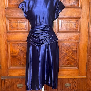 May include: A dark blue satin dress featuring short sleeves, a round neckline, and a gathered, ruched waistline with a scalloped edge detail. The skirt falls to a midi length, and the fabric has a smooth, reflective finish throughout the garment.